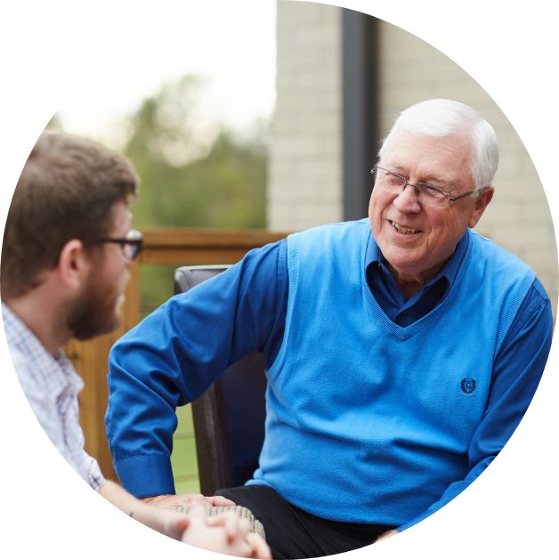 An older man in a blue vest and dress shirt is talking with a younger man outside on a deck